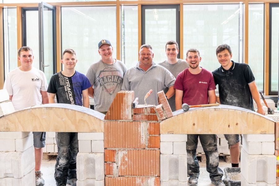 Lehrer mit sechs Schüler stehen hinter einem Bauobjekt aus Ziegel und Holz.